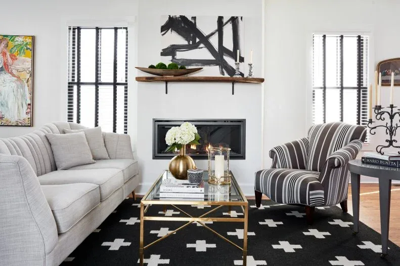 A narrow black-framed fireplace sits below a wood shelf beside black window blinds, a pale sofa, striped chair and dark rug