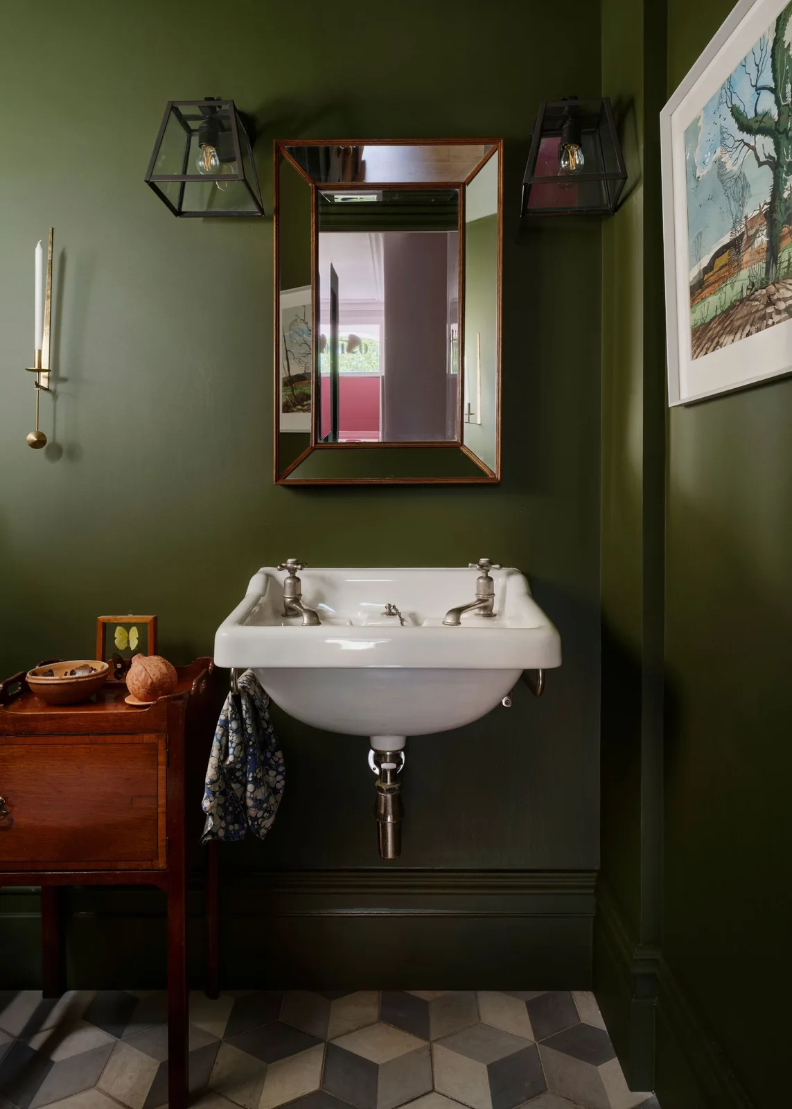 Deep olive walls surround a small white wall-hung basin, a wood chest, a wood-framed mirror and two dark lantern-style lights.