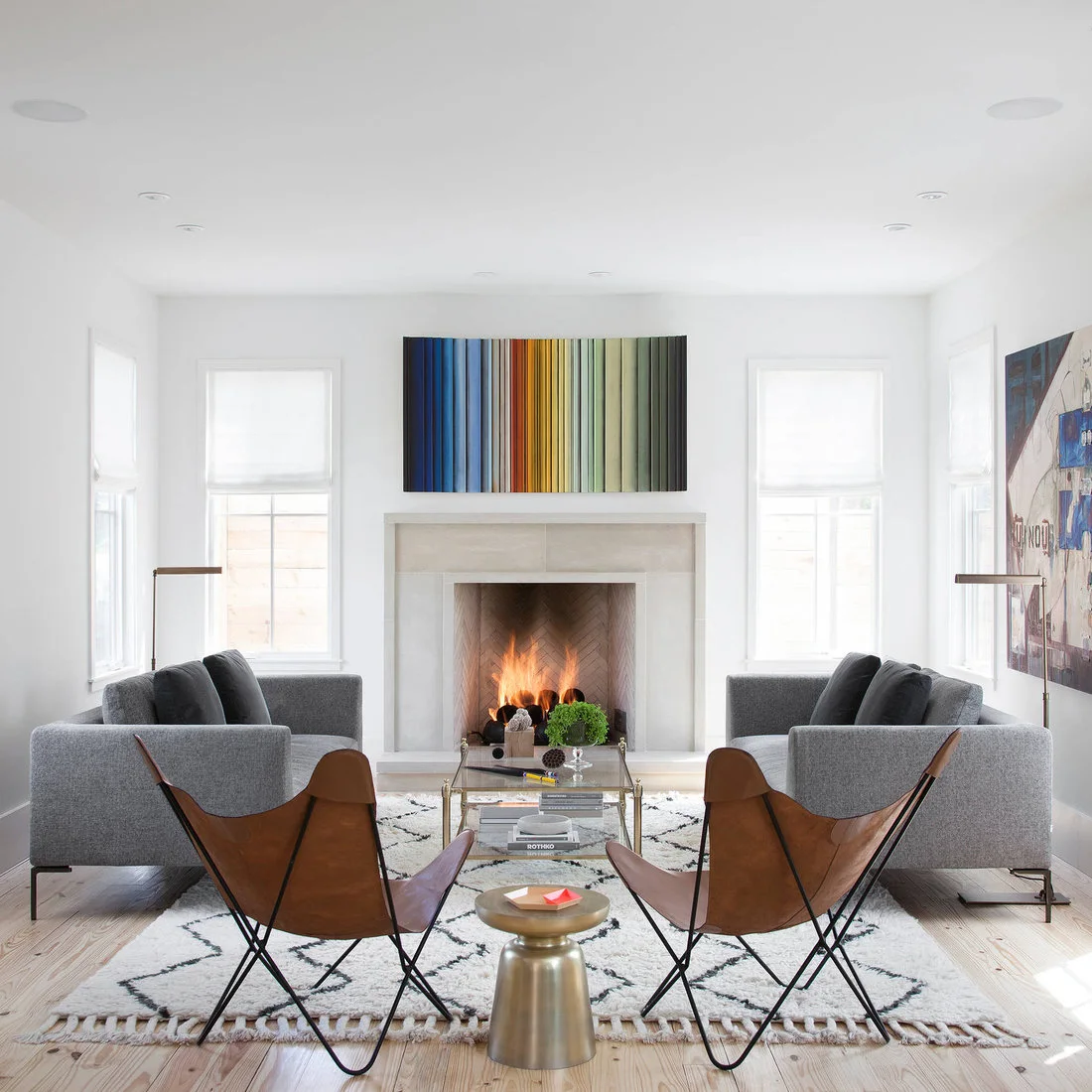 A pale rectangular fireplace stands between windows behind two gray sofas and brown sling chairs, with a colorful striped artwork above and a light geometric rug below.