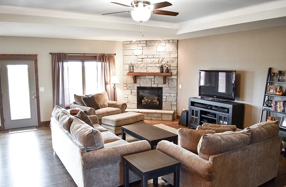 Pale stone corner fireplace with a wood mantel, brown seating and a television on the adjacent wall.