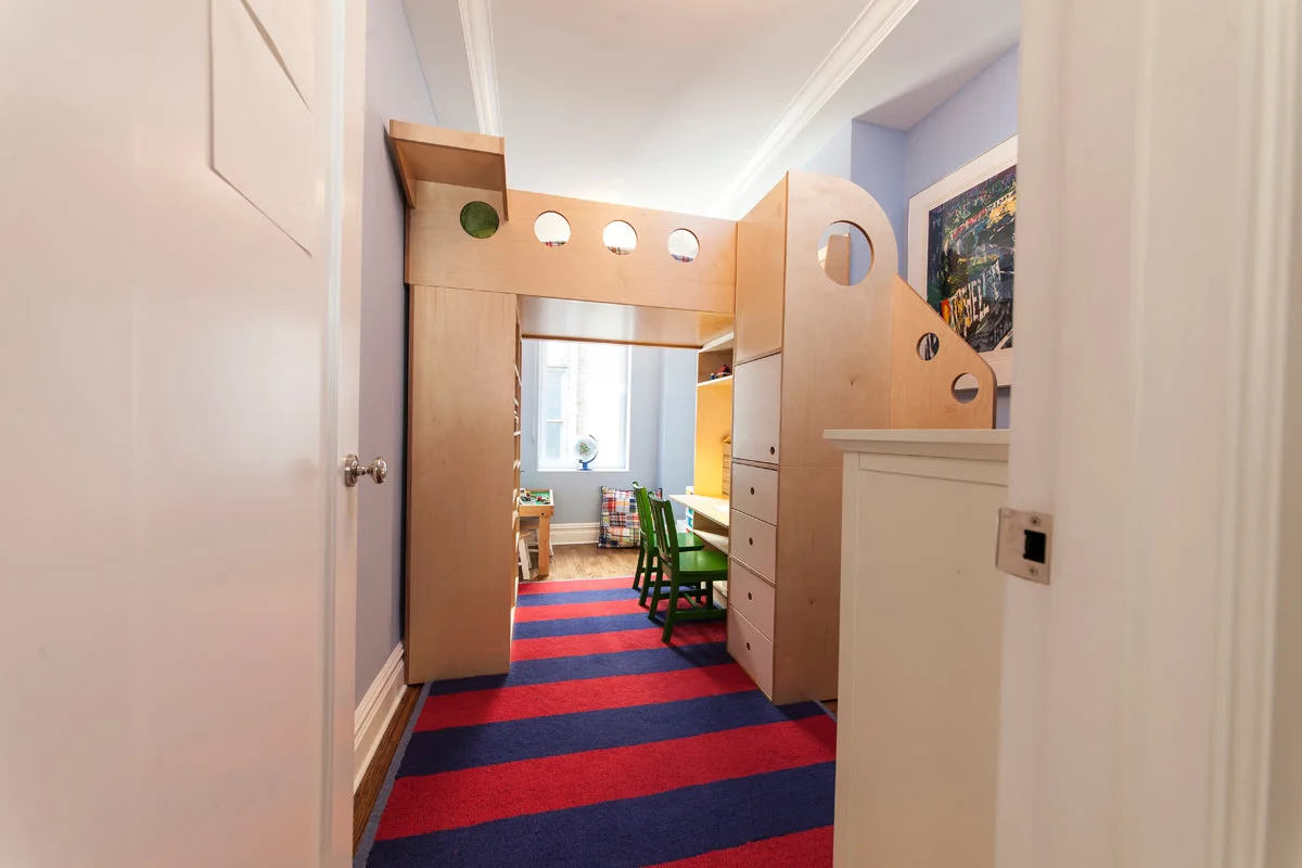 Pale wood loft bed crossing a narrow room with drawer stairs on the right, a desk below and green chairs.