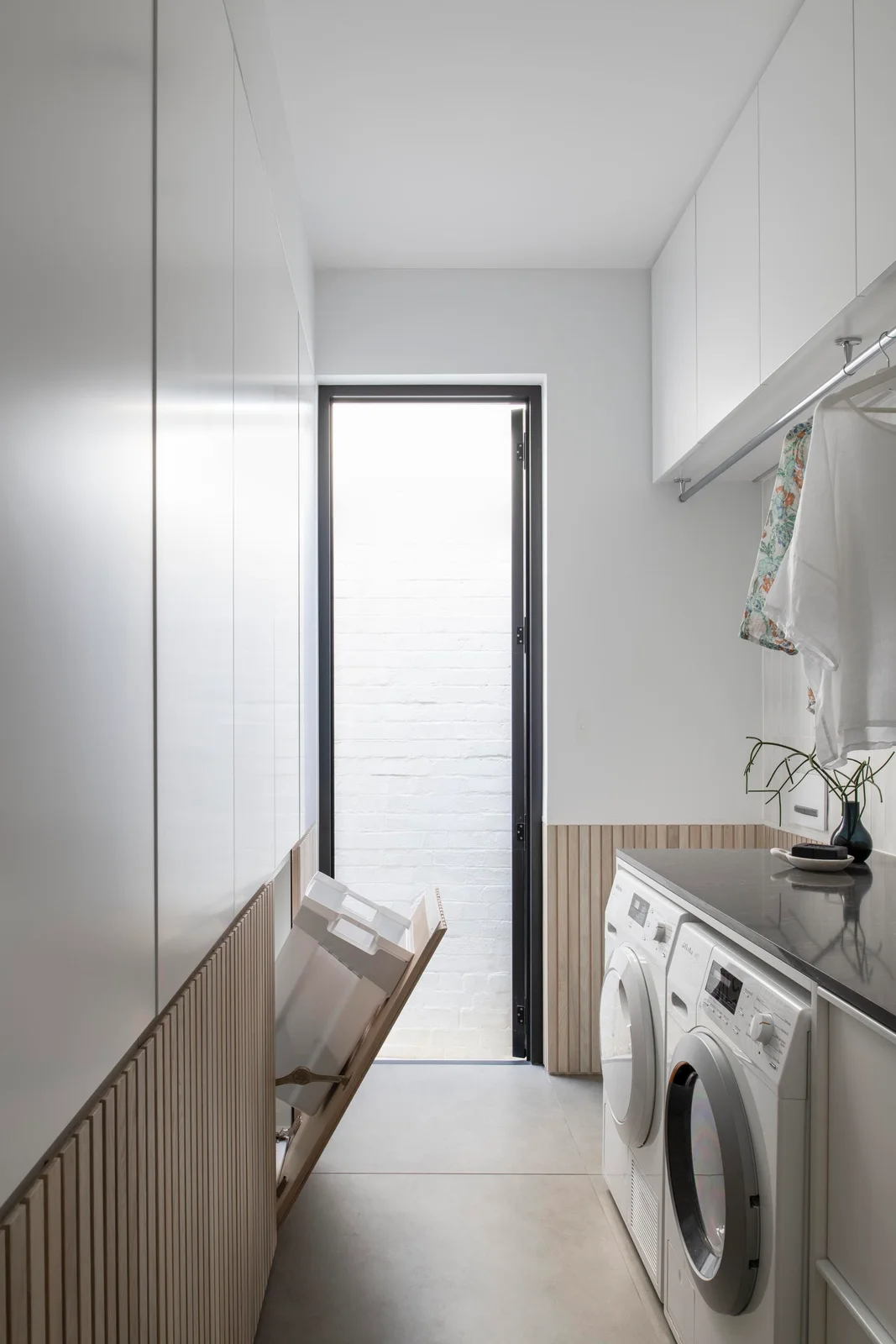 White cupboards with vertical wood detailing face side-by-side laundry machines, with a tilt-out hamper and a glazed door at the end of the narrow room.
