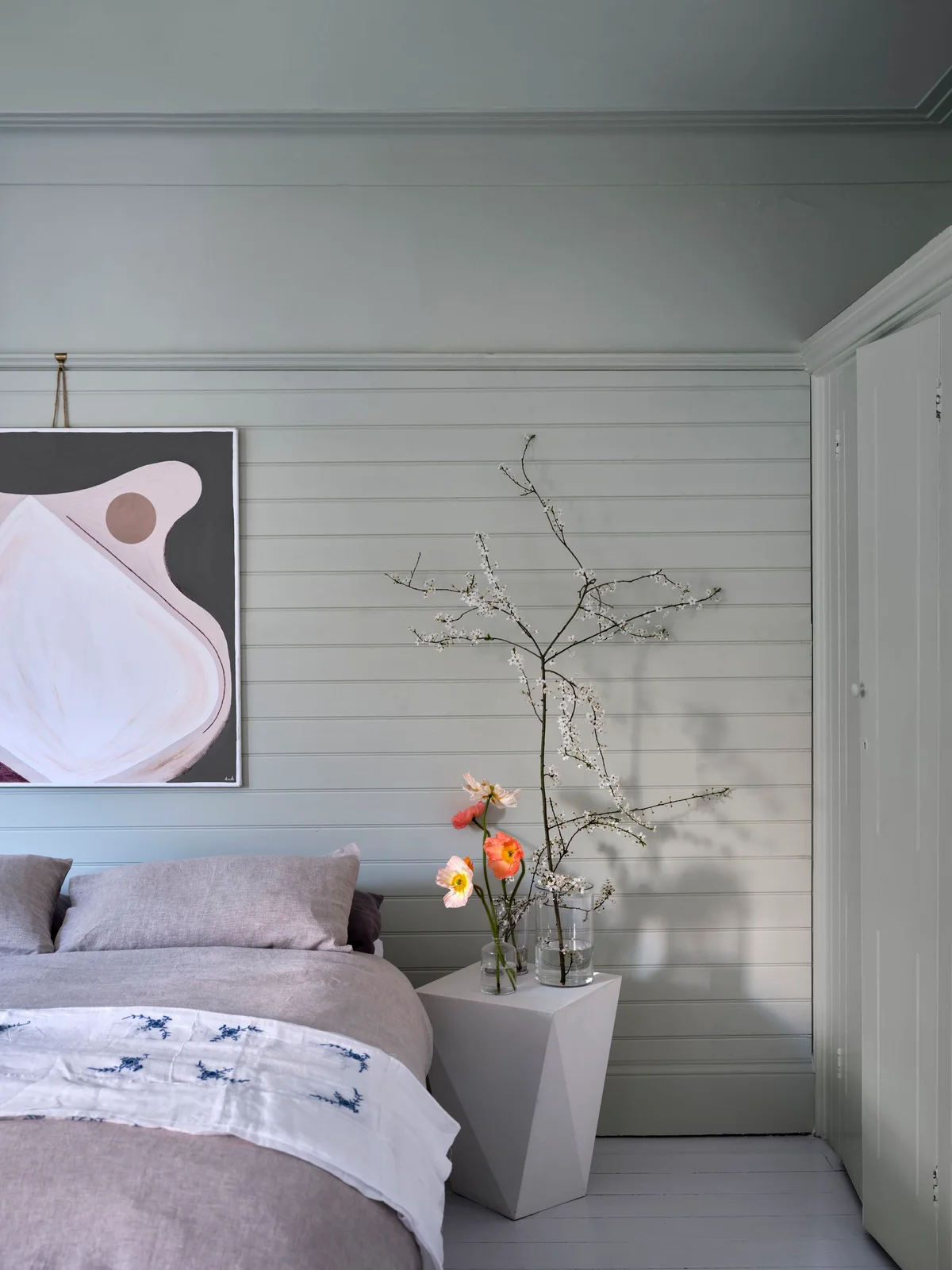 Gray-green horizontal wall boards rise behind pale bedding, a framed abstract print and a small angular white bedside table holding cut flowers and branches.