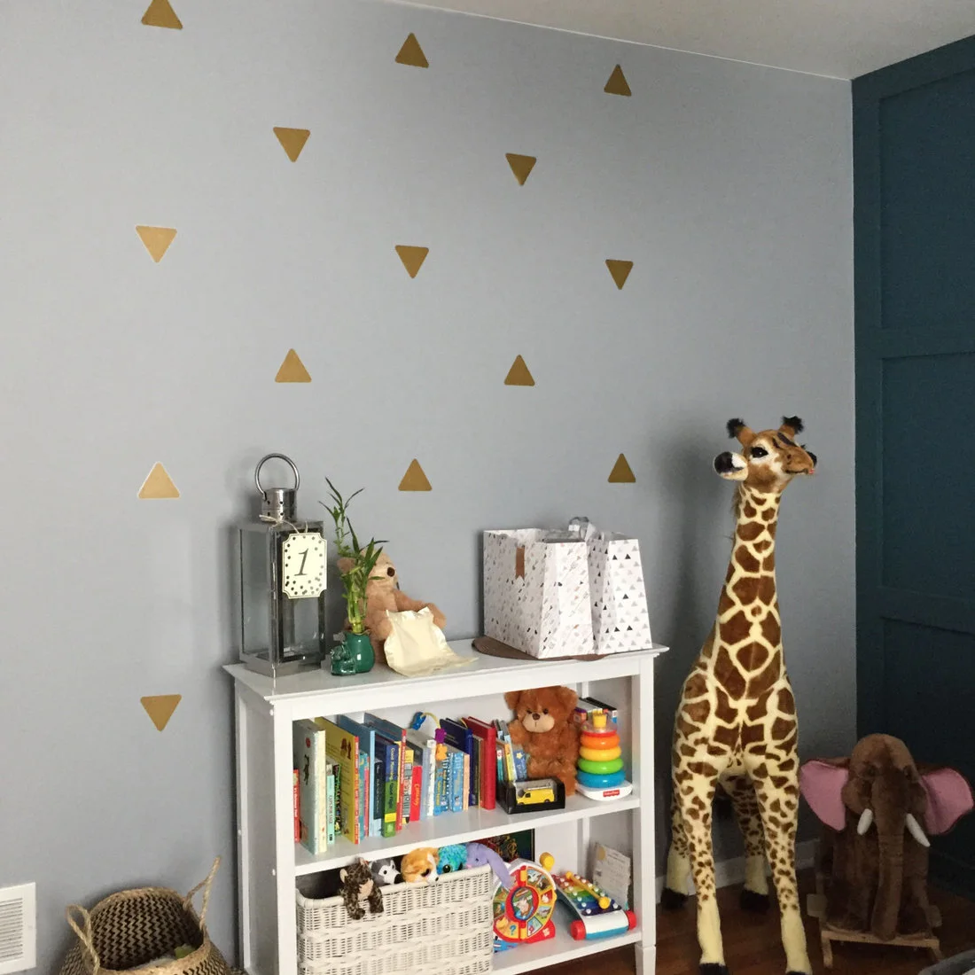 A low white cube bookcase holds books, baskets and toys against a gray wall with gold triangles beside a tall toy giraffe.