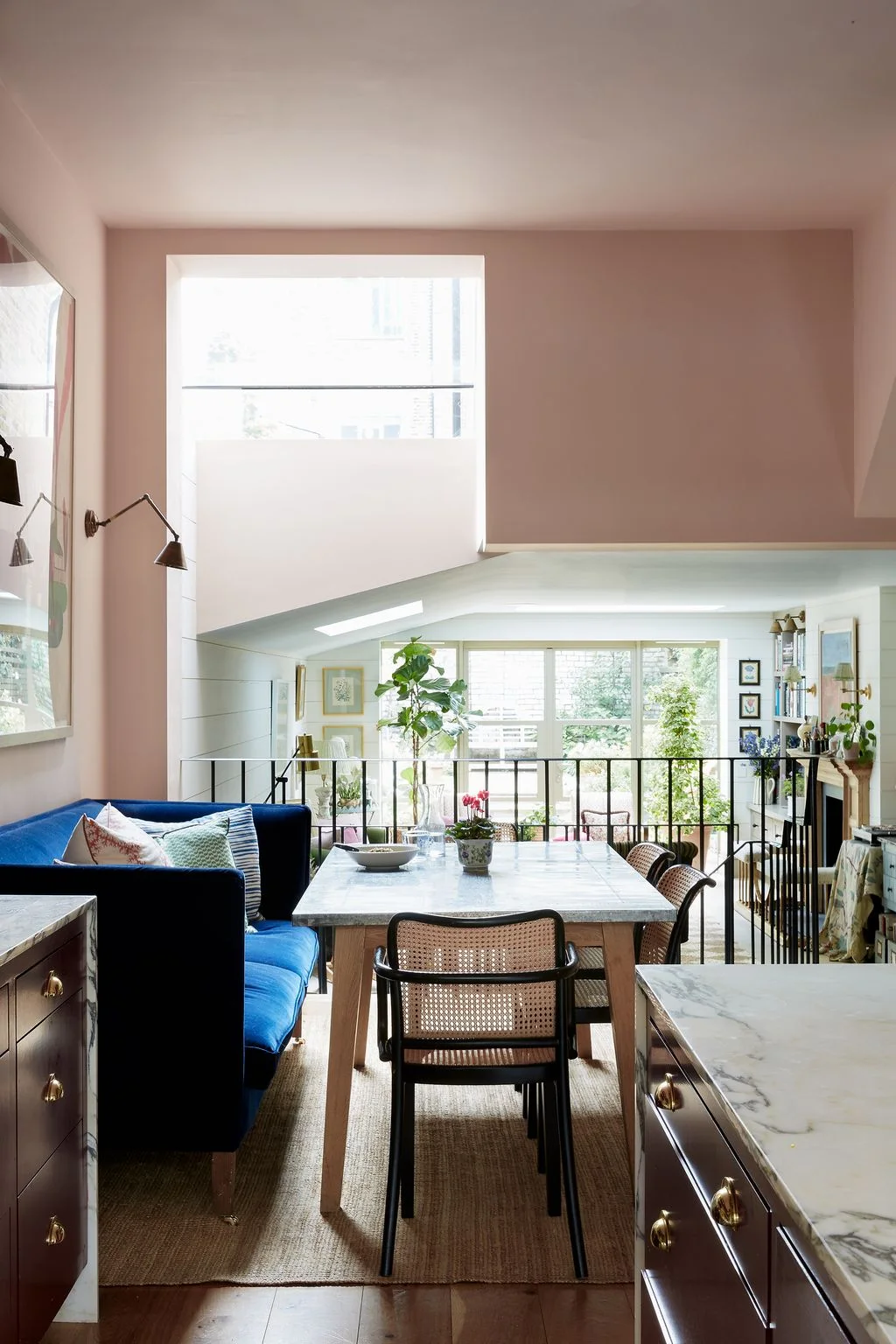 A dining table and blue banquette sit beside kitchen counters above a lower sitting room with tall windows.
