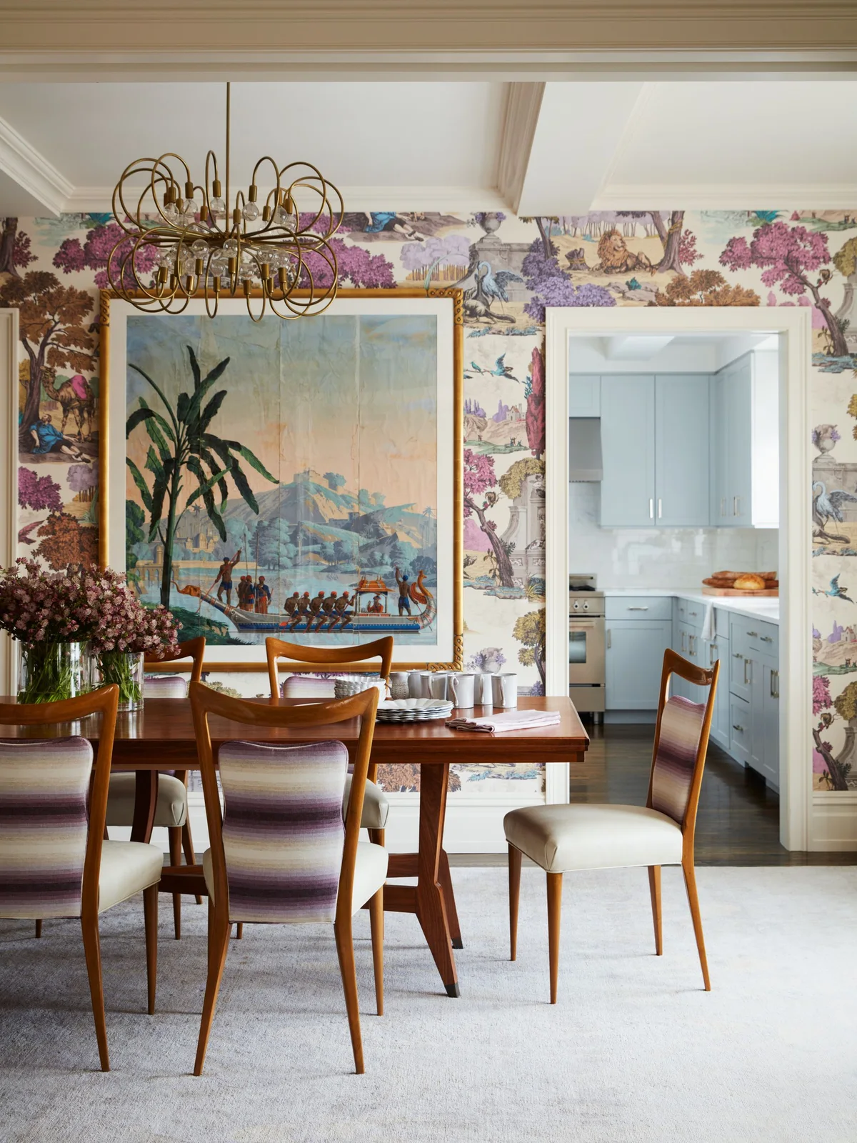 Illustrated wallpaper surrounds a wooden dining table and striped chair backs beneath a large framed landscape and a curved metal chandelier