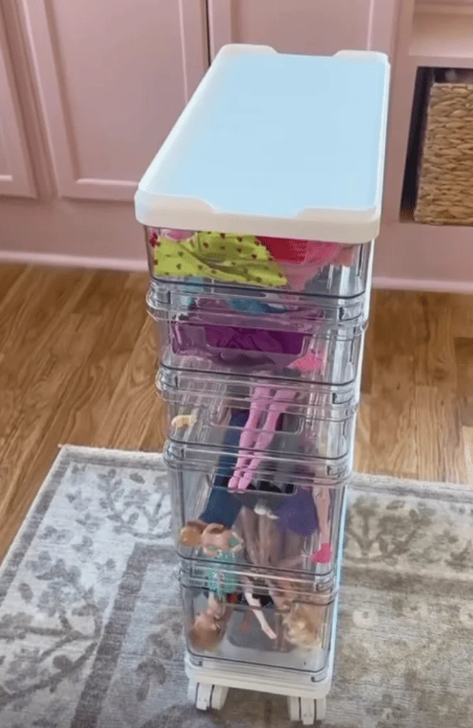Slim clear drawer cart on casters with shallow upper drawers and deeper lower drawers holding dolls and accessories.