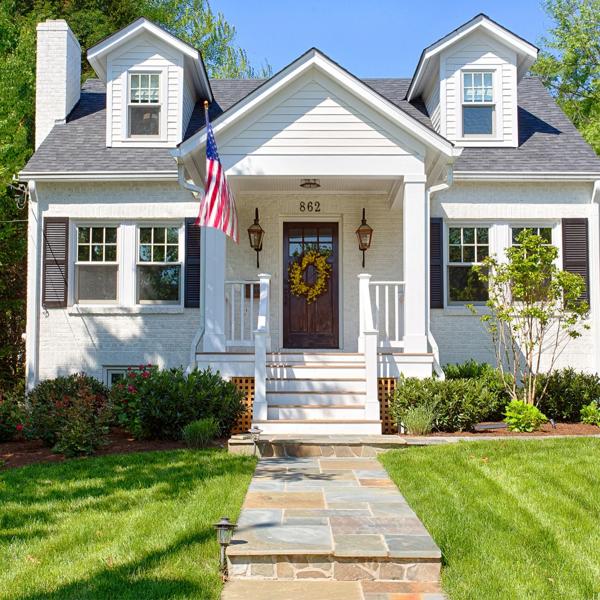 A white Cape-style house has two roof dormers and a small gabled porch with white posts, railings and a straight path.