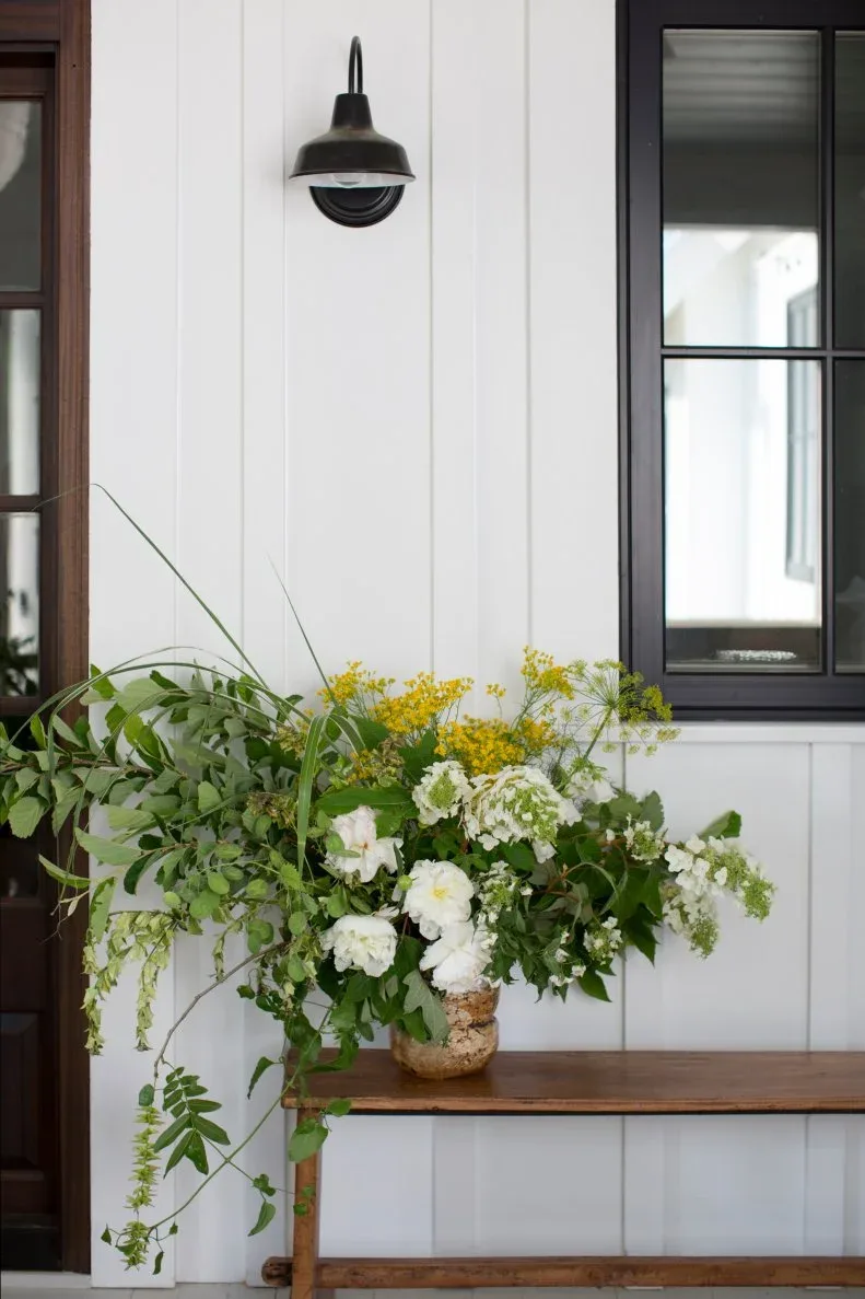Wood bench beneath black-framed porch window with loose white and yellow flower arrangement
