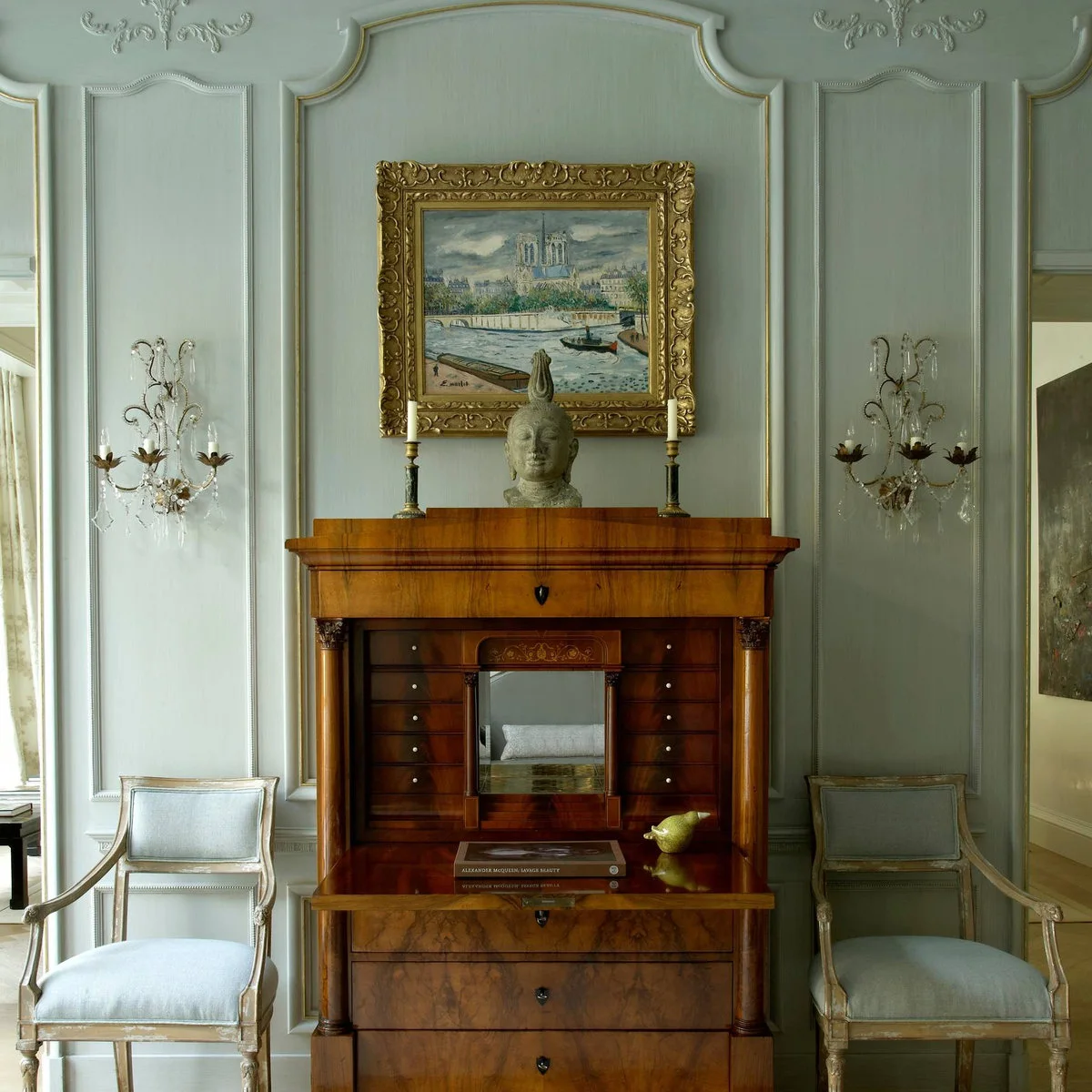 Warm wood cabinet and framed artwork against pale gray paneled walls with light upholstered chairs.
