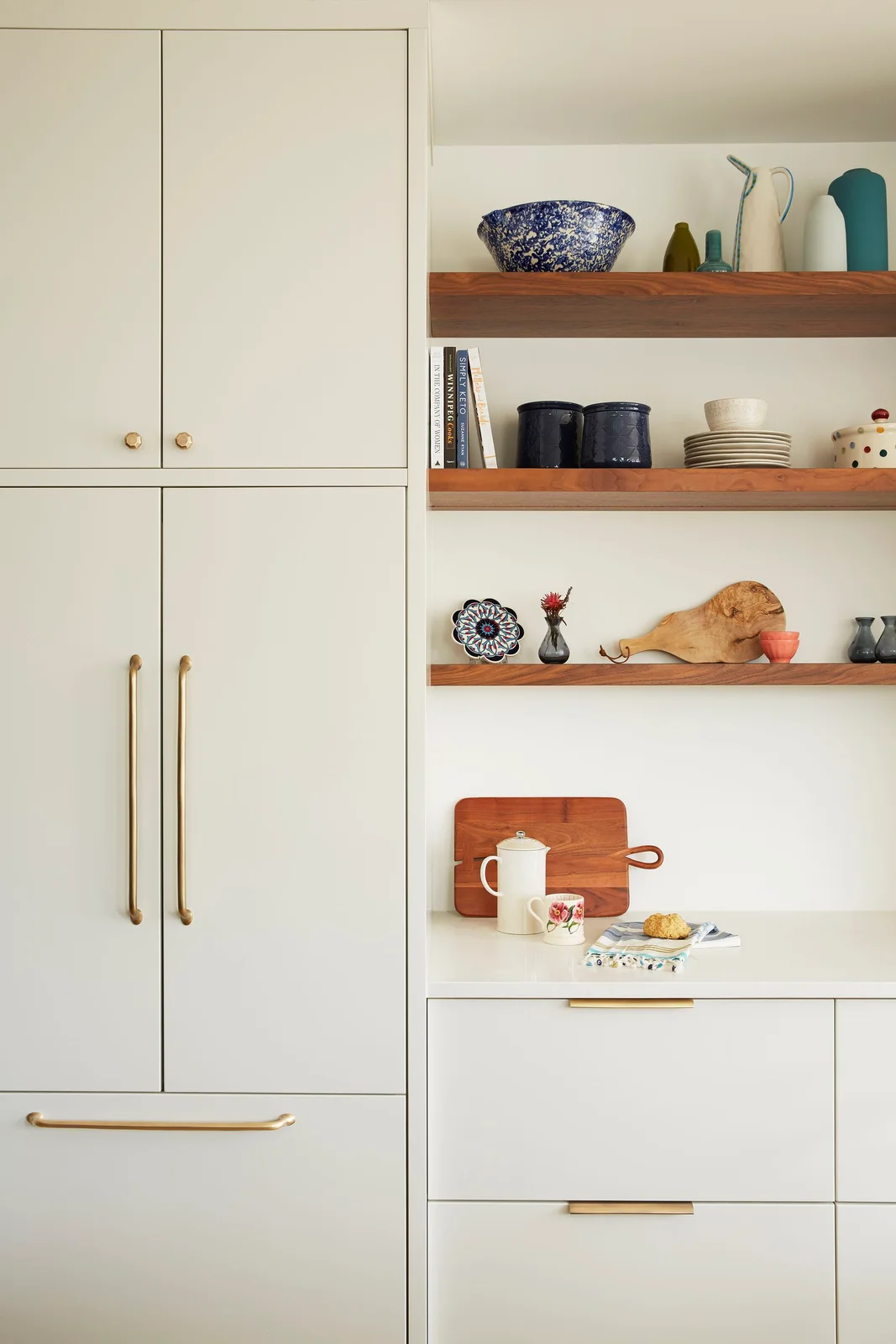 Pale flat-front kitchen cabinets with gold-toned pulls stand beside wooden shelves holding dishes above a light counter