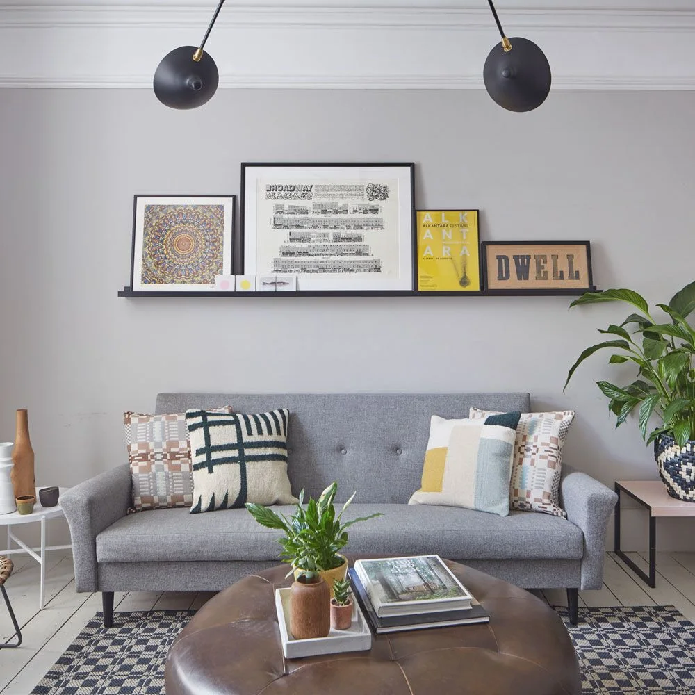 A gray sofa with patterned cushions sits beneath a narrow picture ledge against a pale gray wall beside a leafy plant and round brown ottoman