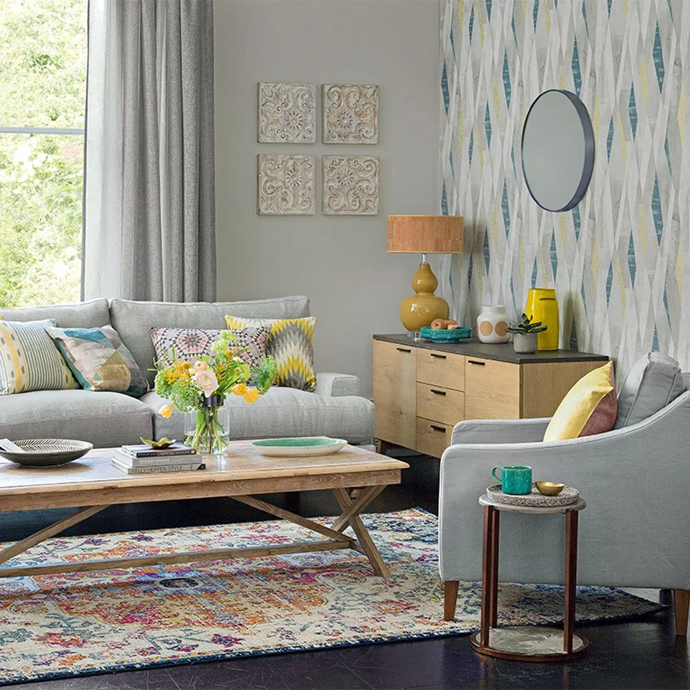 Blue-and-gray geometric wallpaper sits behind a wooden cabinet with a round mirror beside gray sofas and a patterned rug