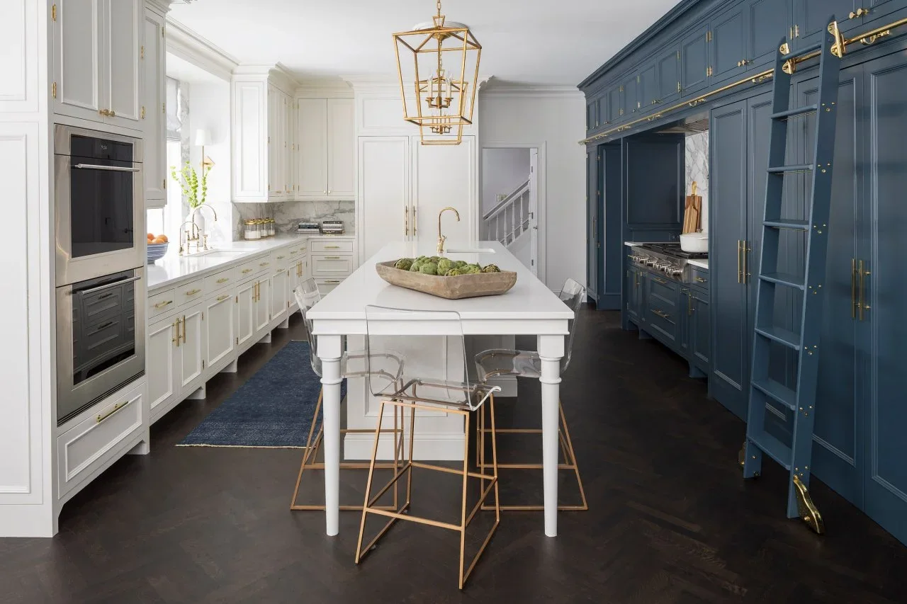 A navy cooking and storage wall faces white kitchen cupboards across a white island with clear stools and gold-colored lights.