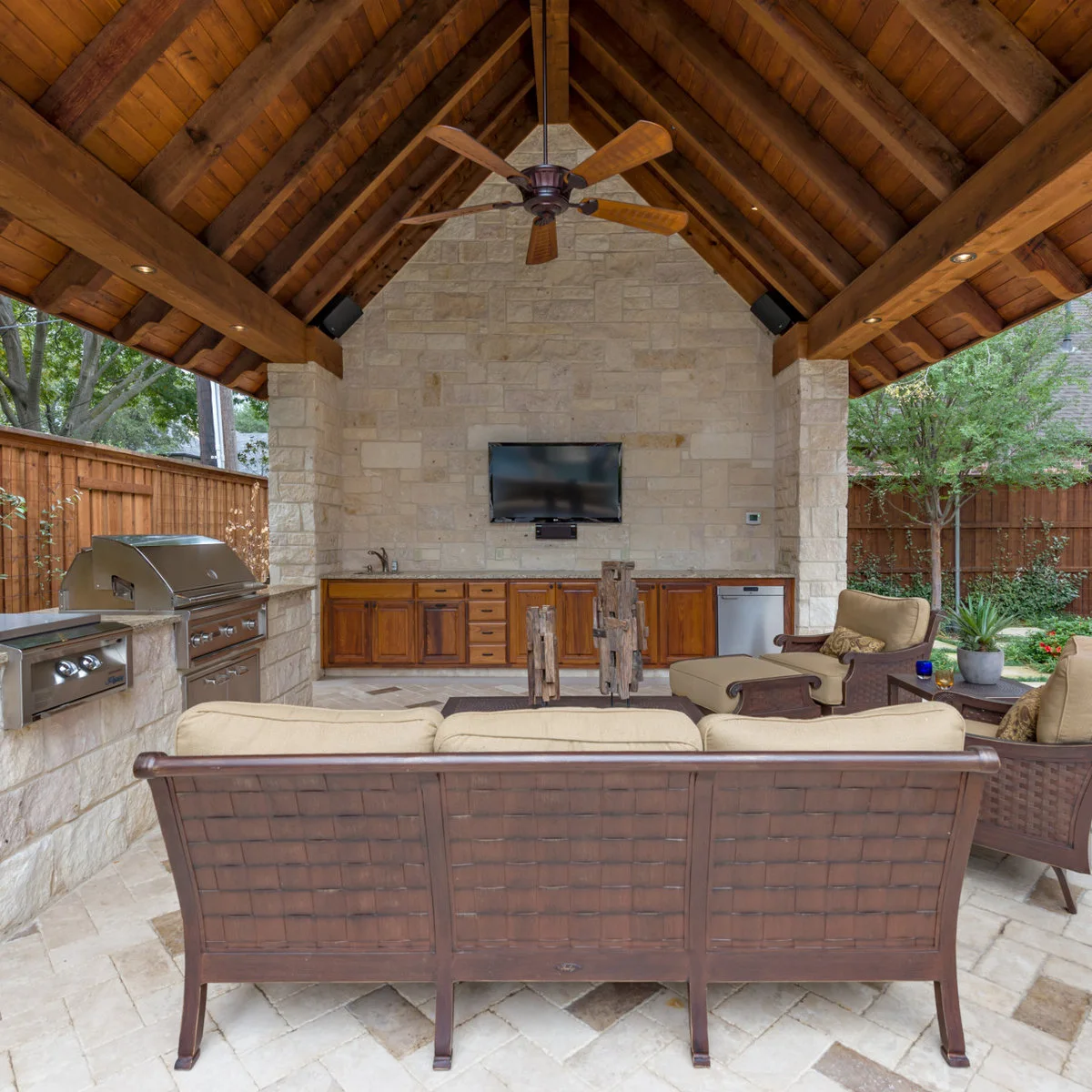 Outdoor pavilion with grill along one side, sink and wood cabinetry at the back and cushioned seating in front