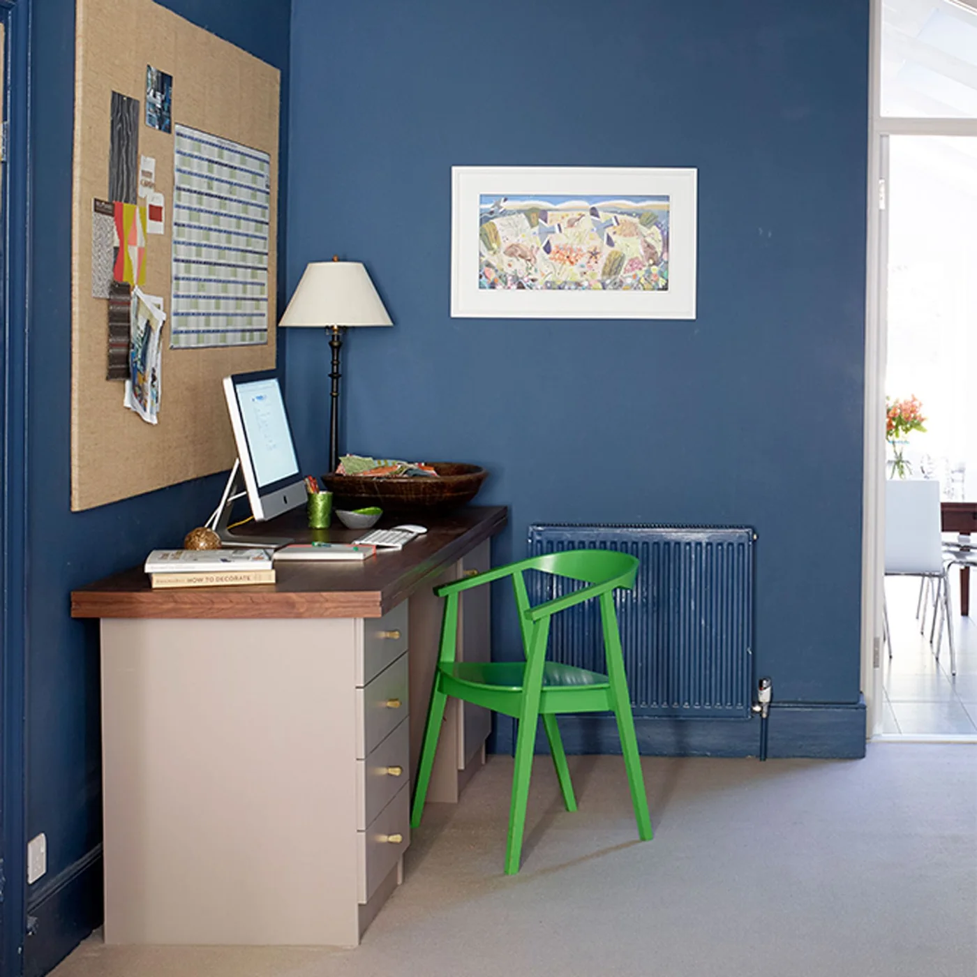 A bright green wooden chair sits at a dark-topped desk with pale drawers against blue walls beneath a large noticeboard