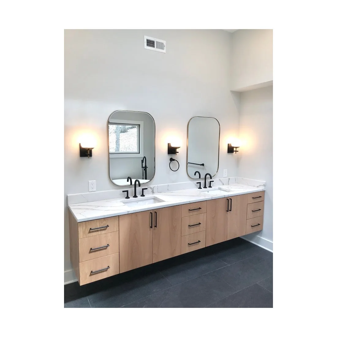 A long pale wood double vanity floats above a dark tiled floor, with black taps and handles, rounded rectangular mirrors and three wall lights.