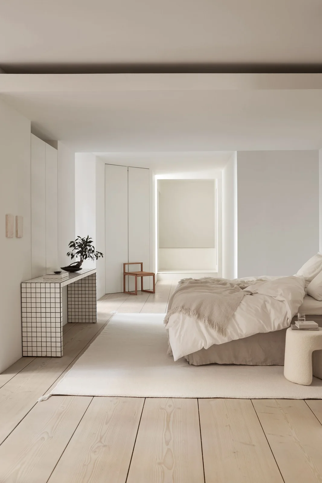 A white bedroom has broad pale floorboards, simple fitted cupboards, a tile-grid console, a pale bed and a large light rug beside a small wood chair.