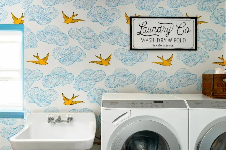 White laundry appliances beside pale sink under wallpaper with yellow birds and blue cloud-like patterns and a framed laundry sign
