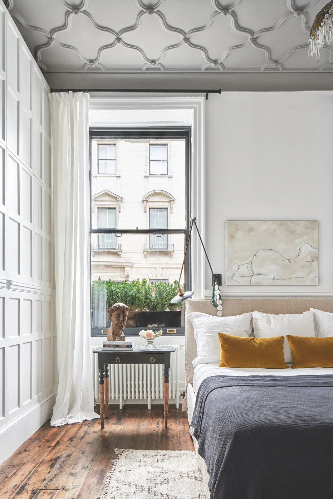 A white paneled bedroom has a patterned pale ceiling, dark gray bed covering, mustard cushions and a tall window with light curtains