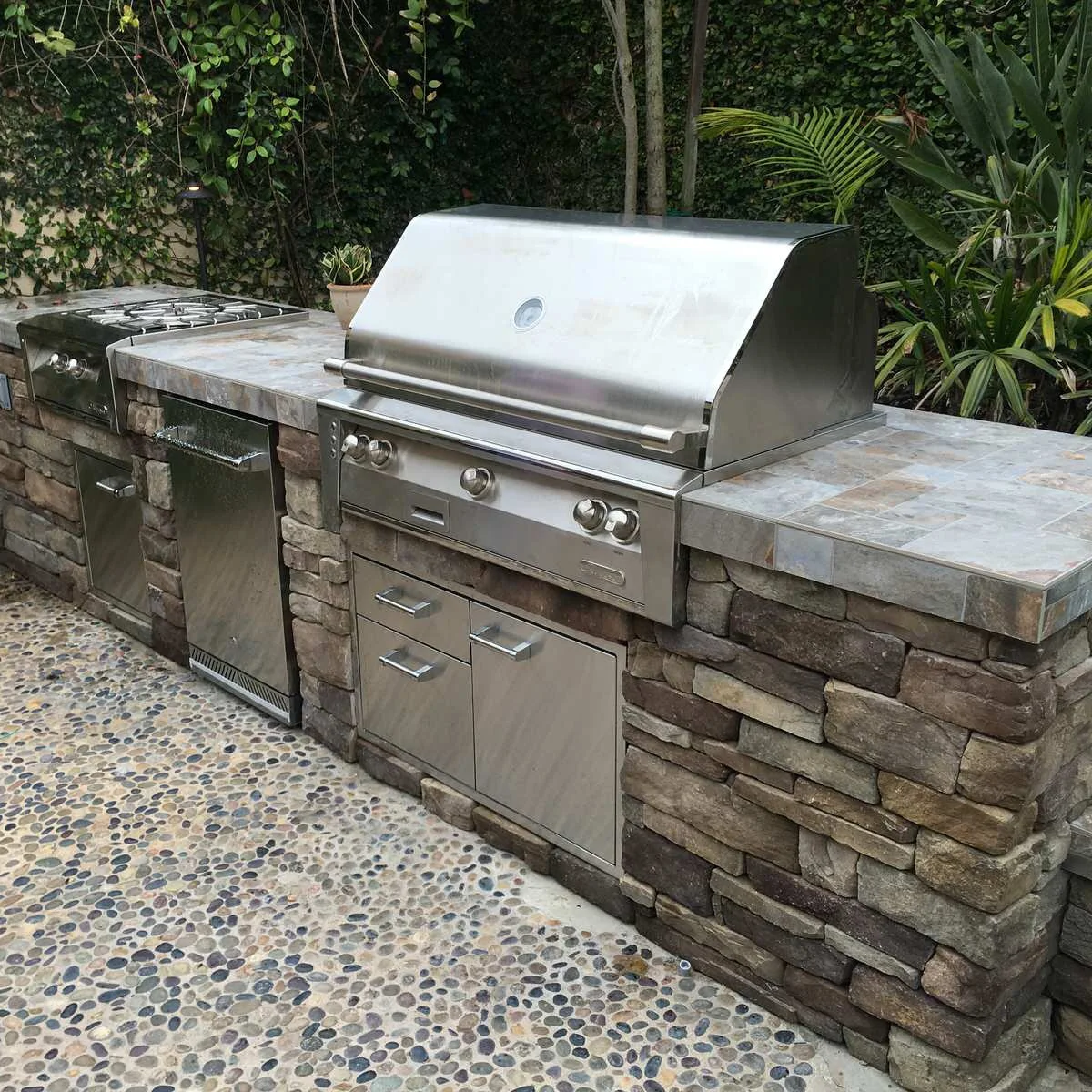 Outdoor grill run with tiled counters, stone-faced bases, small refrigerator and pebble-pattern paving