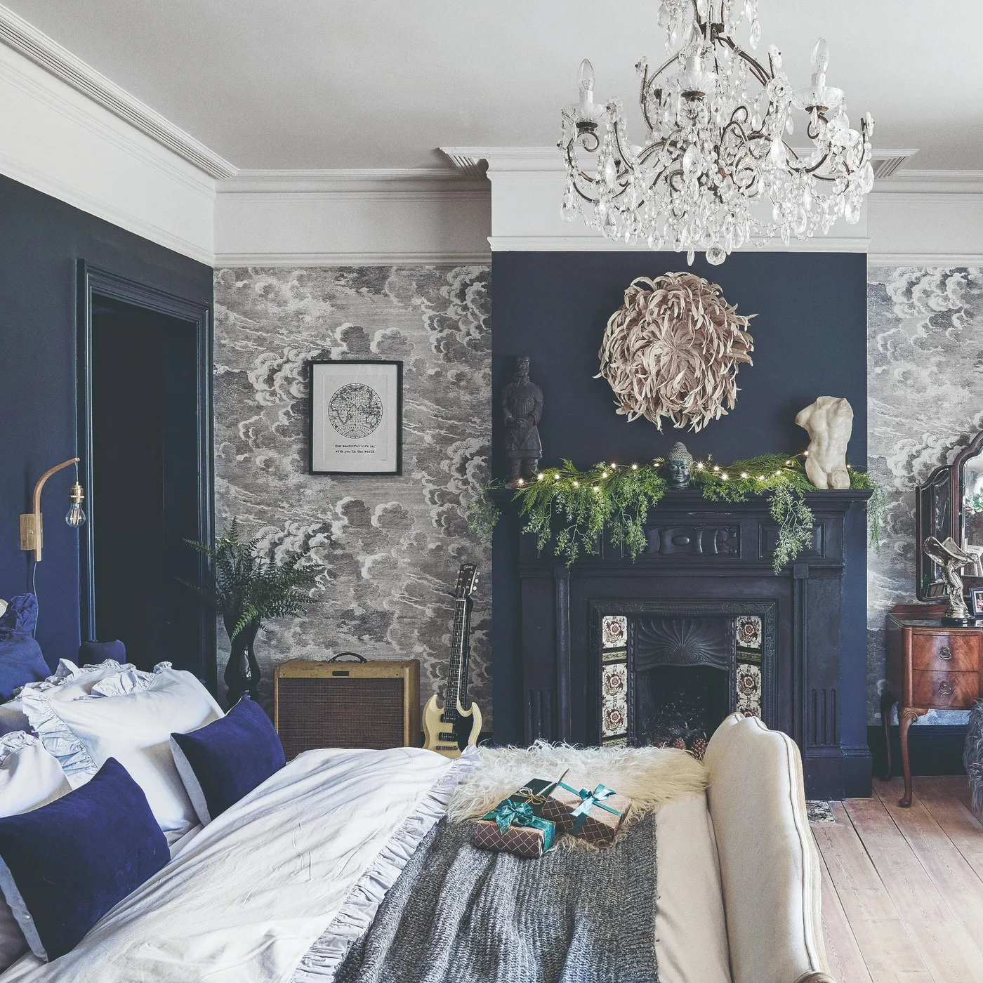 Navy bedroom walls and fireplace surround with patterned wallpaper panels, white cornicing, a chandelier, and a wooden floor.