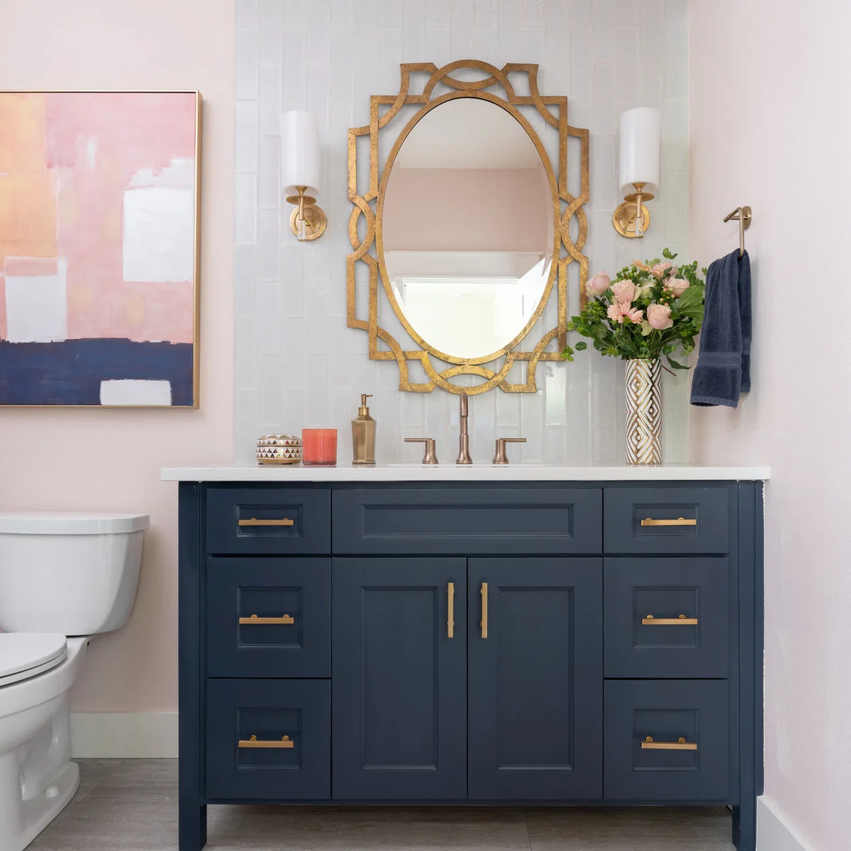 Navy vanity beneath gold-colored mirrors with pink walls, pink accessories and a vase of flowers.