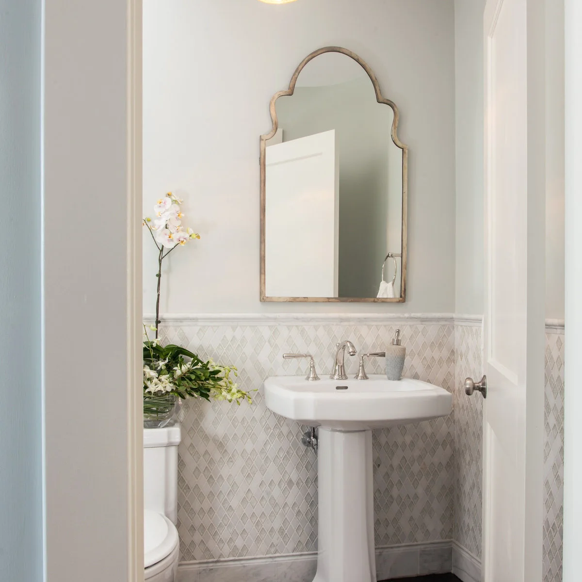 A white pedestal sink sits against pale diamond-patterned tiles below a tall shaped mirror beside an inward-opening door.