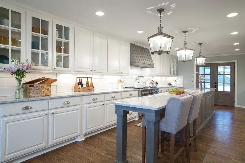 A long dark-front island with a pale counter stands beside white framed cabinets and glass upper doors beneath simple lantern-shaped pendant lights