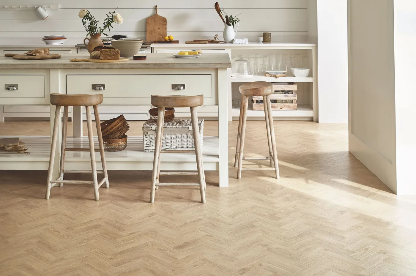 Pale kitchen with open-backed island storage, three wooden stools, baskets and herringbone-patterned floor.