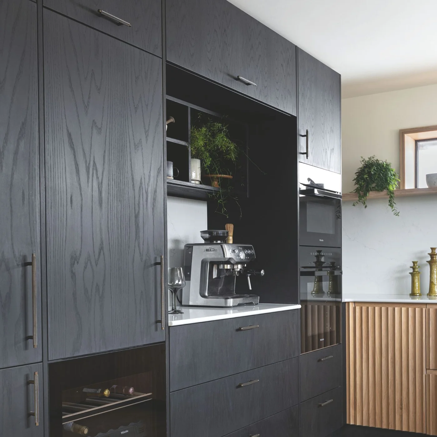 Black-stained oak tall cabinets surround an open coffee niche and stacked ovens, beside a natural oak fluted cabinet run.