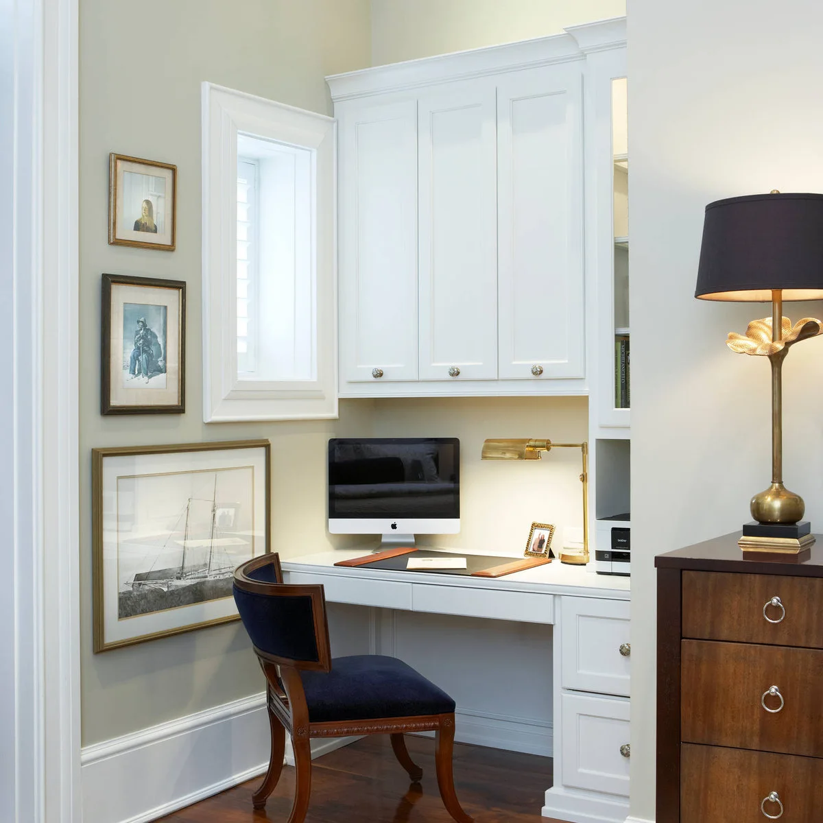 White compact desk beneath paneled cabinets with a dark chair and brass-colored adjustable lamp