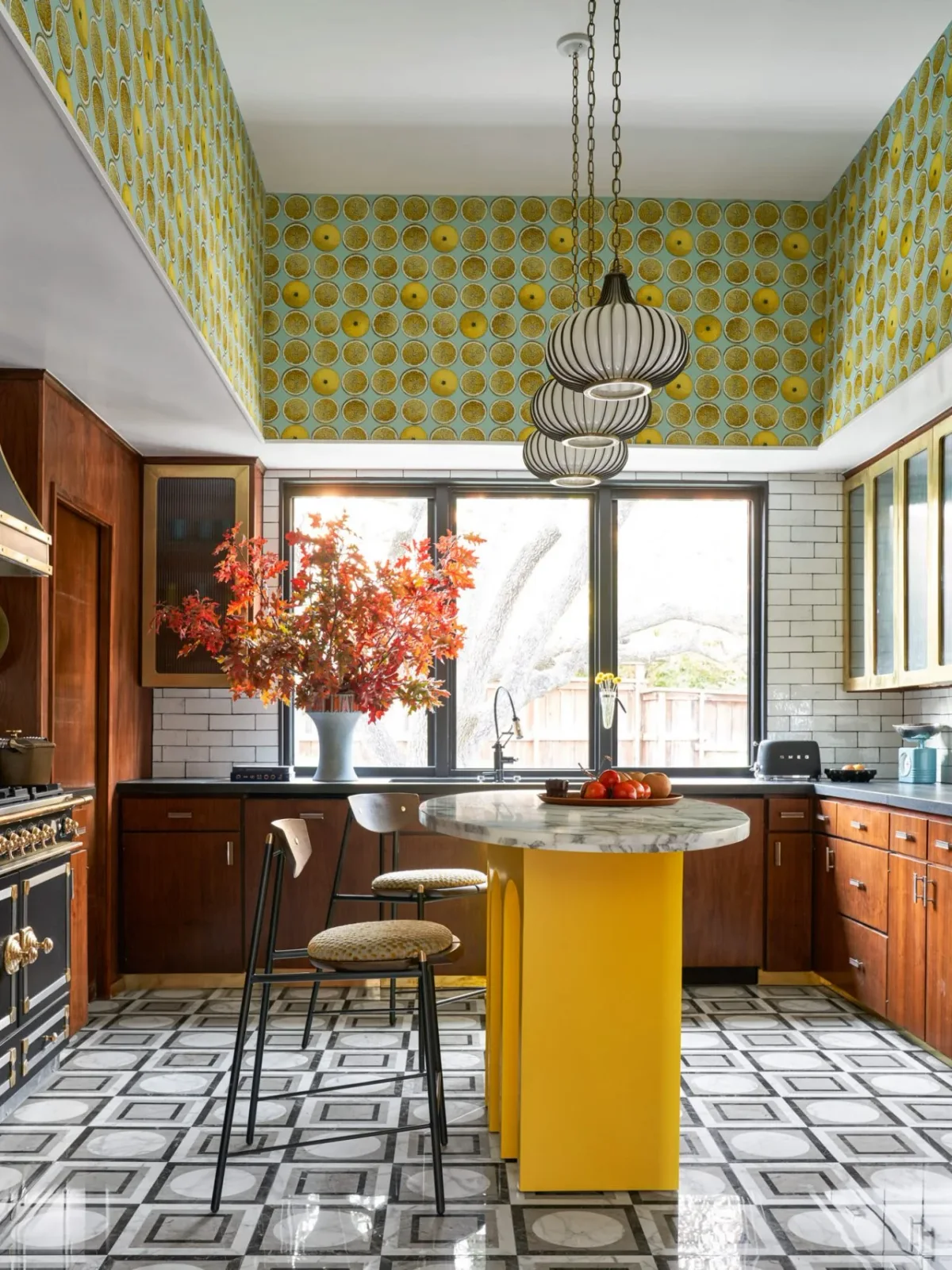 Warm wood cabinets surround a yellow island, with lemon-patterned upper walls, white tile and a black-and-white patterned floor.
