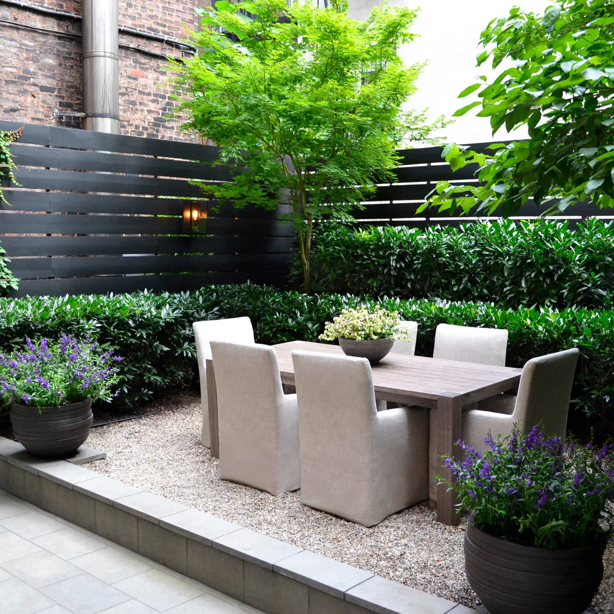 Four pale upholstered outdoor chairs surround a wood table on a gravel terrace enclosed by planting and dark horizontal fencing.