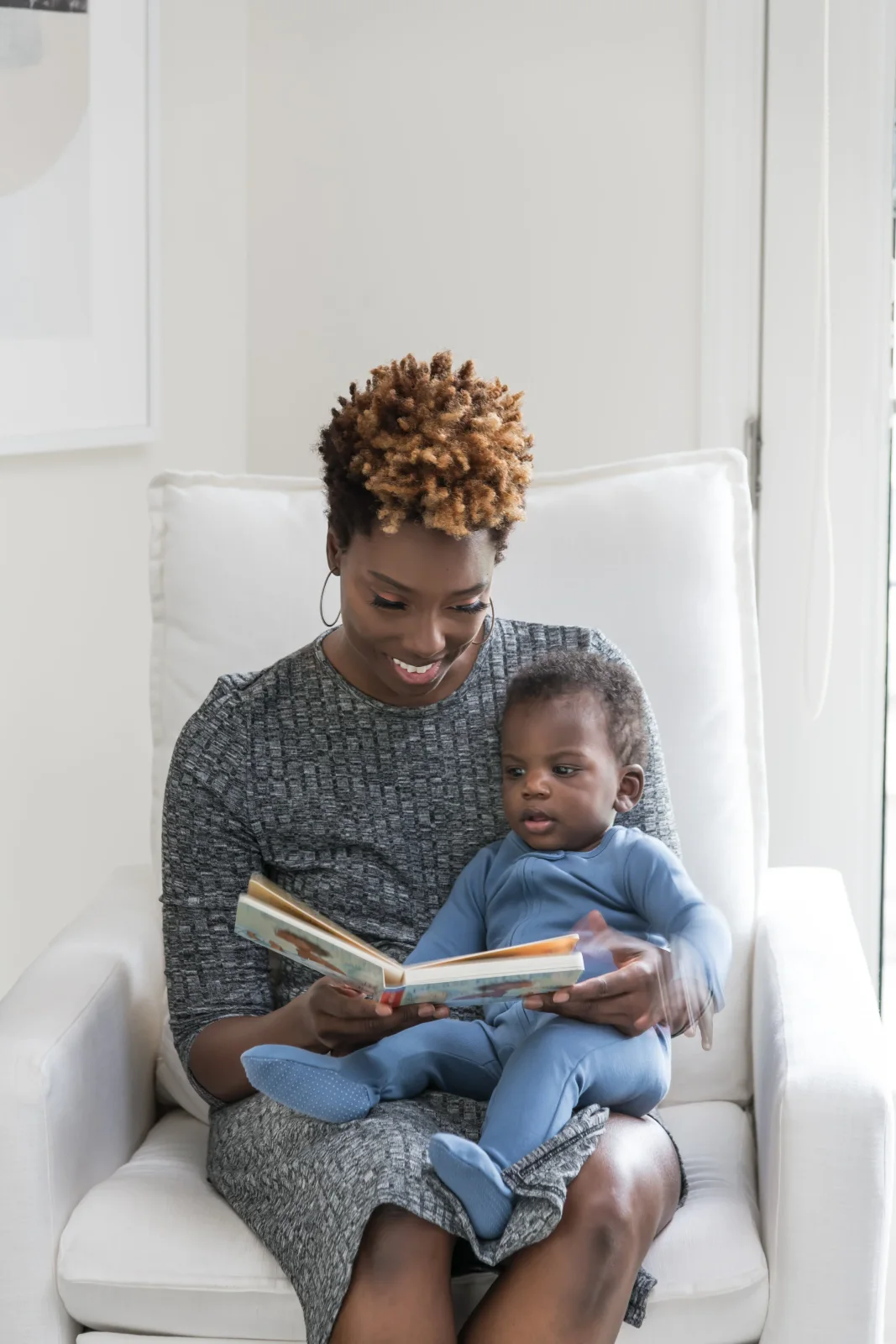 An adult sits in a pale nursery chair holding a child and an open book beside a light wall and window
