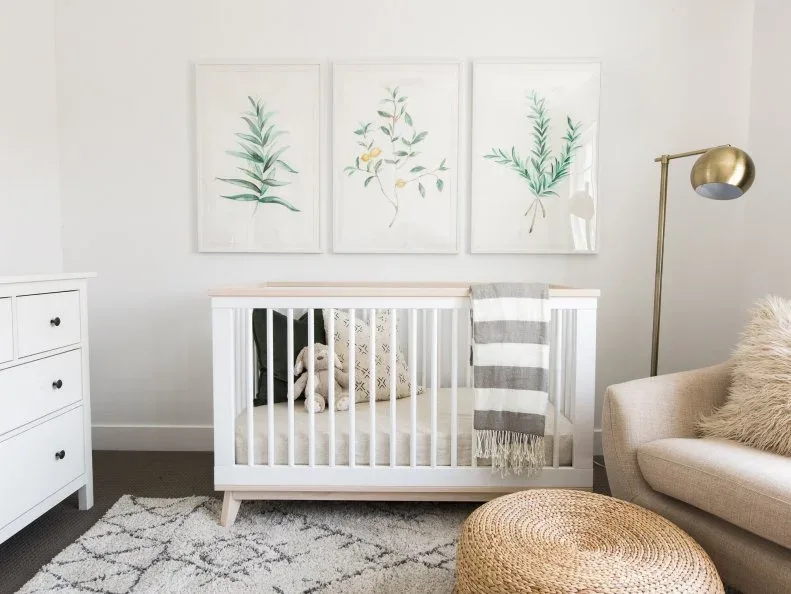 Pale nursery with white crib, three botanical pictures, adult sofa, woven round pouf, striped throw and slender floor lamp.