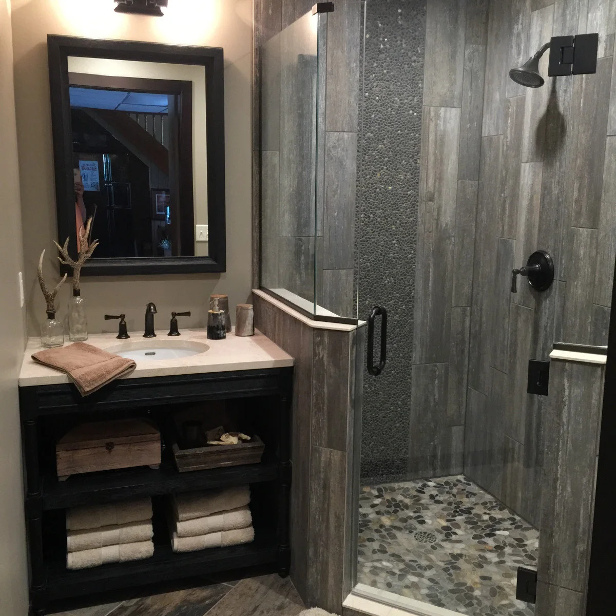 A gray wood-grain tiled shower with a narrow pebble stripe, matching pebble floor, black fittings and a pale-capped partition.