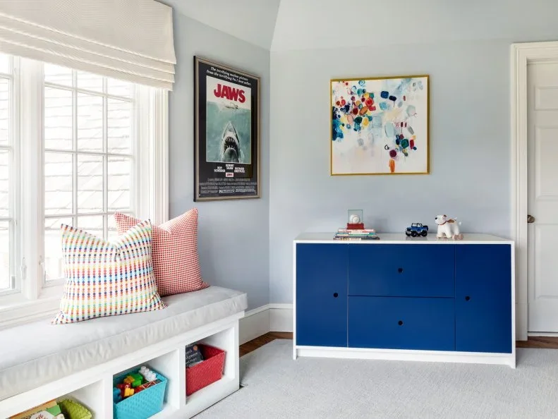 A pale blue room has a low white cabinet with dark blue doors and drawers beside a cushioned white window bench with colorful bins beneath.