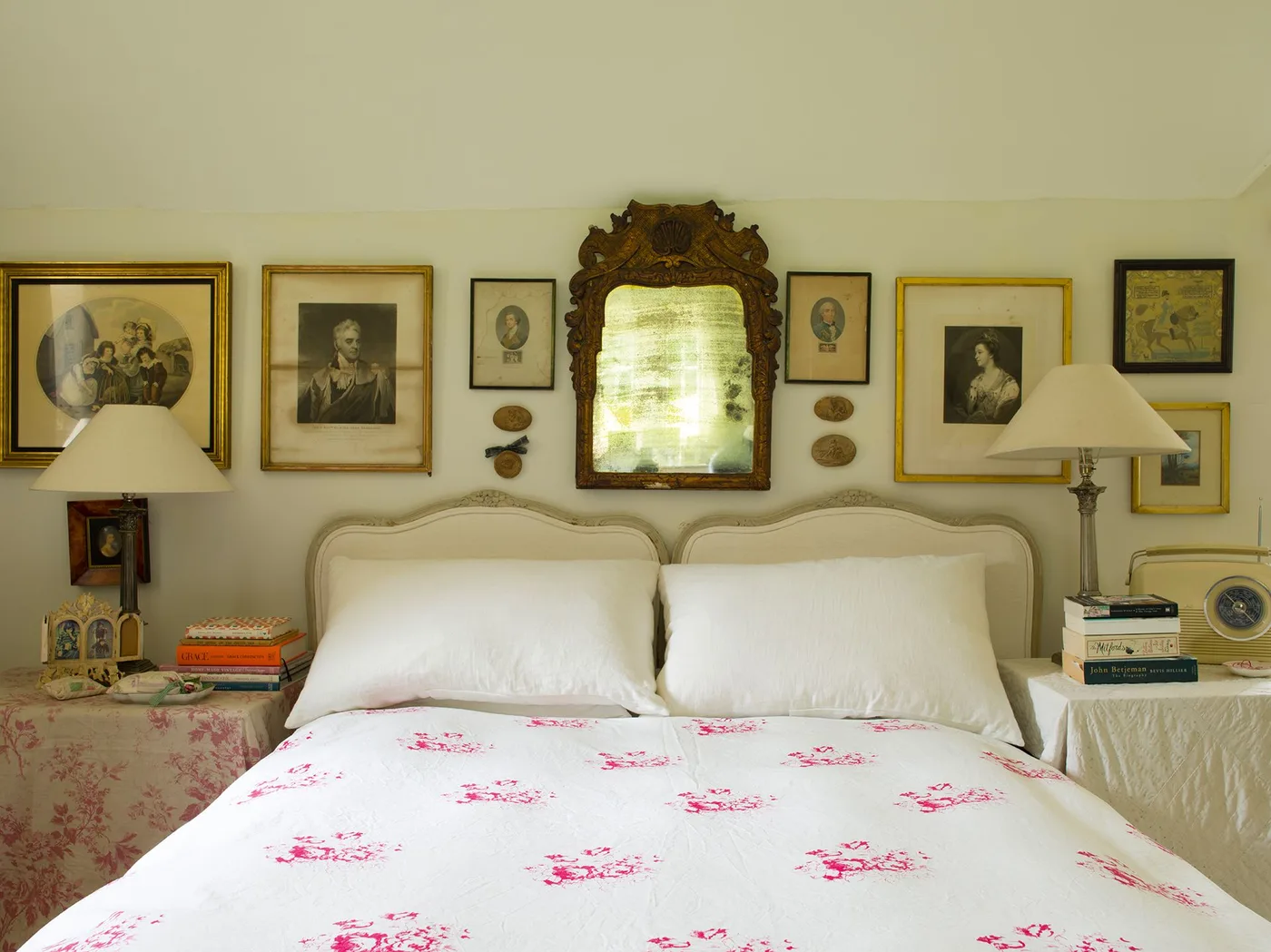 Traditional bedroom with pale twin-shaped headboard sections, an ornate mirror, framed portraits and pink floral bedding.