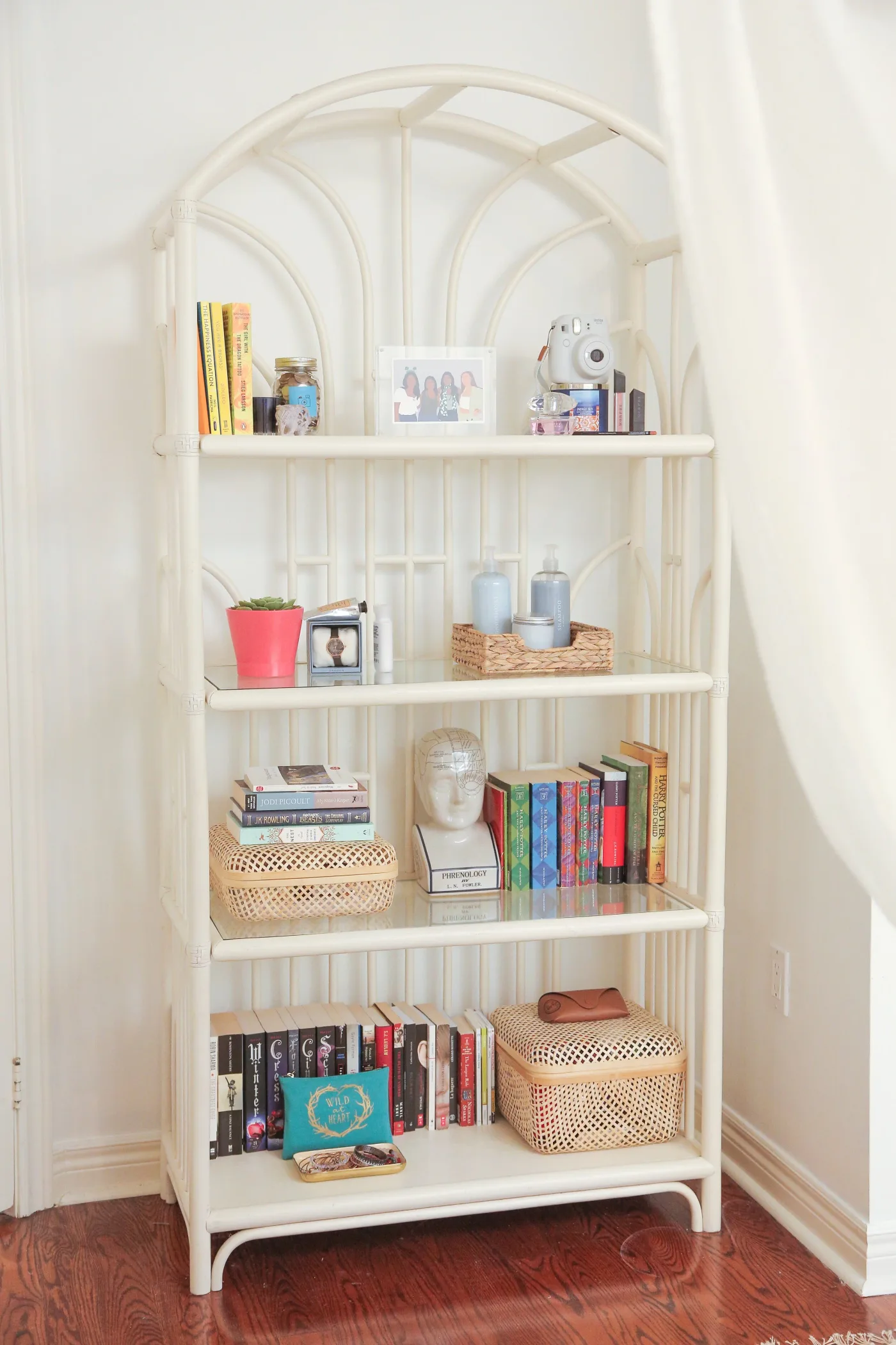 Pale arched open bookcase in a student bedroom with upright books, small woven boxes, framed photograph and camera.