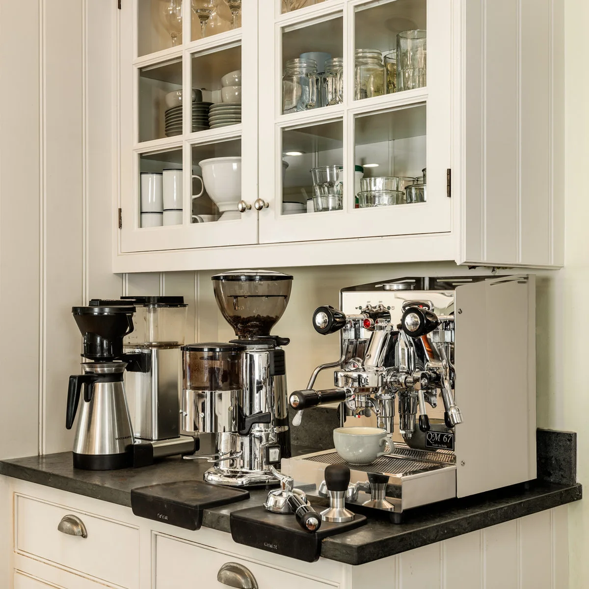 Espresso machine and grinders on a dark counter beneath white glazed cupboards