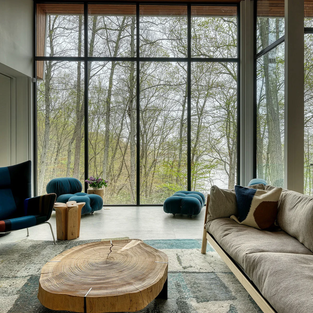 Gray sofa and rug with teal seating, a broad wood coffee table and tall windows overlooking trees.