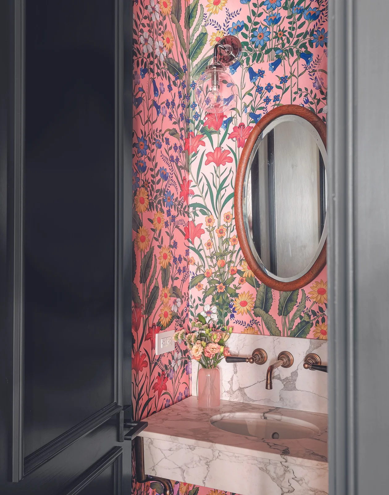 A dark paneled door opens toward pink floral wallpaper, a round wood-framed mirror, clear glass wall light and pale veined basin counter.