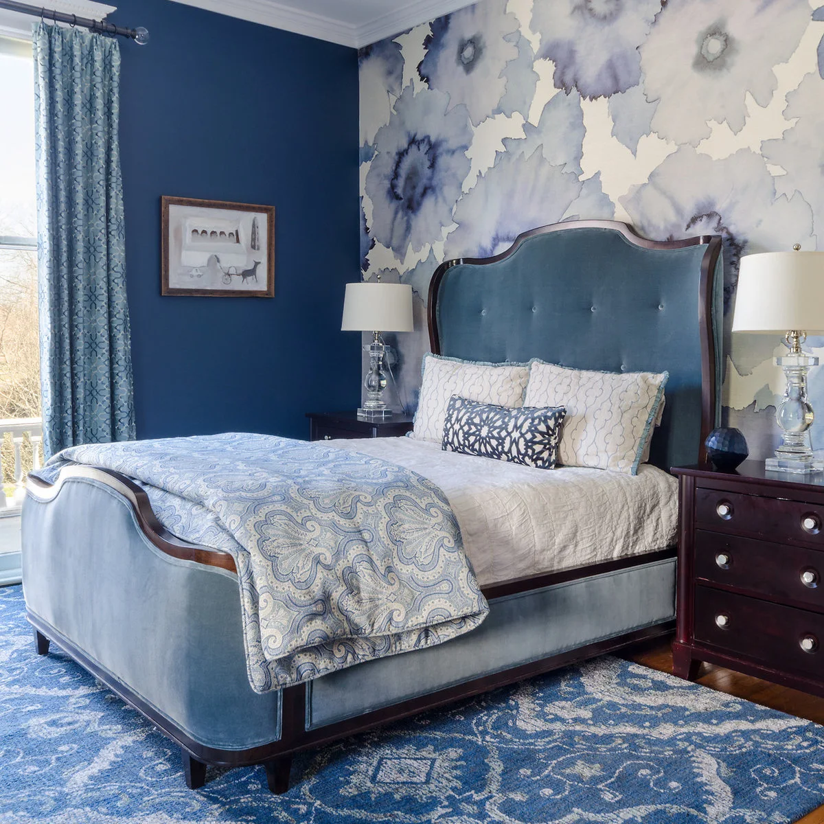 Blue upholstered sleigh bed with floral wallpaper, blue curtains and a dark wooden bedside chest.