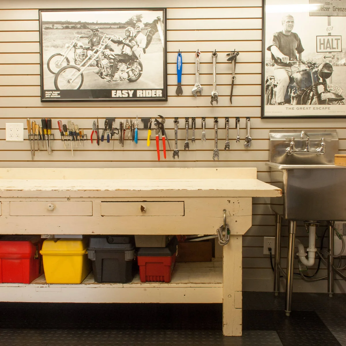 White garage workbench with colorful boxes beneath, hand tools on a slat wall and a utility sink alongside.