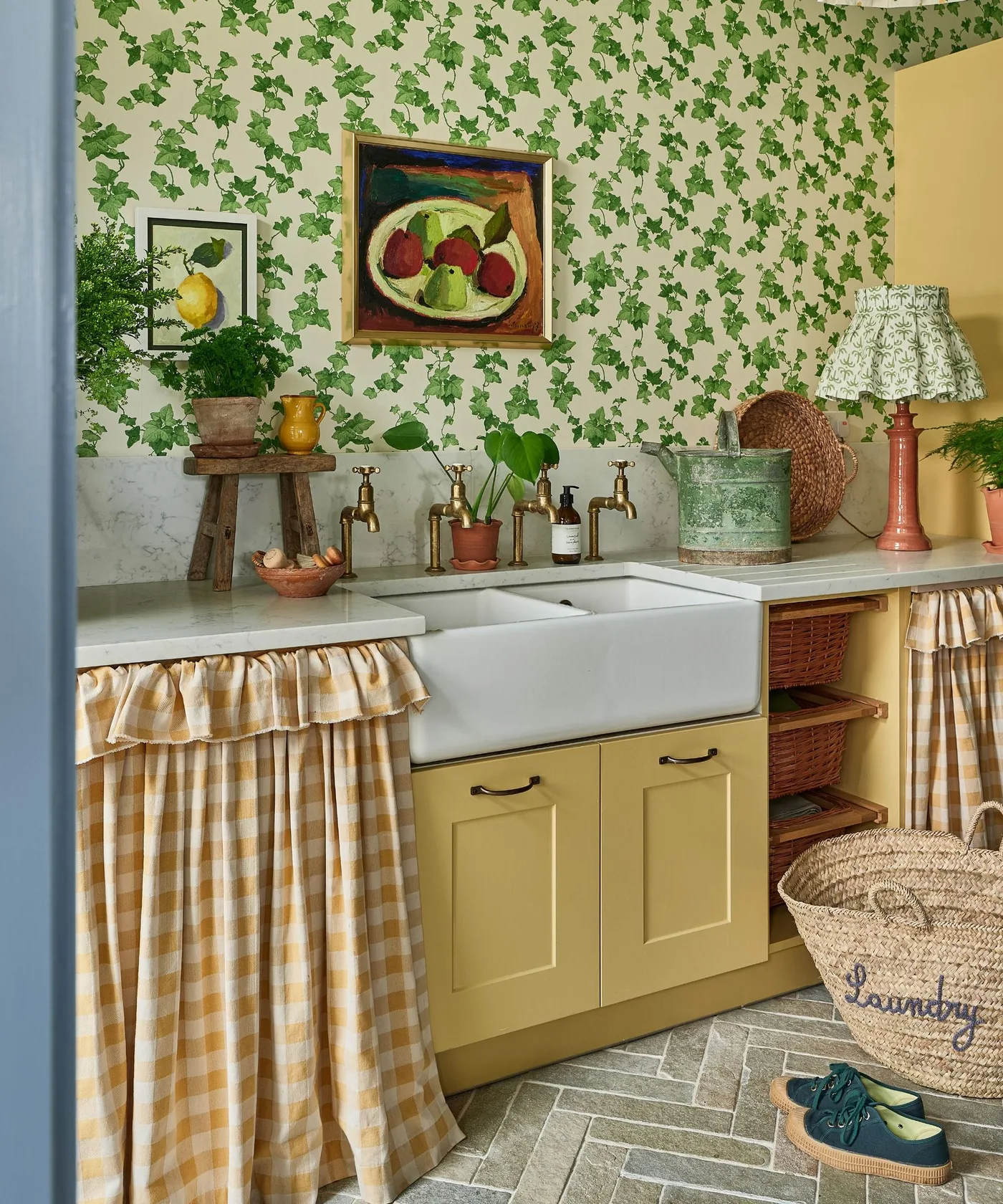 Yellow utility cupboards with ivy wallpaper, white apron-front sink, checked fabric curtains, baskets and framed pictures