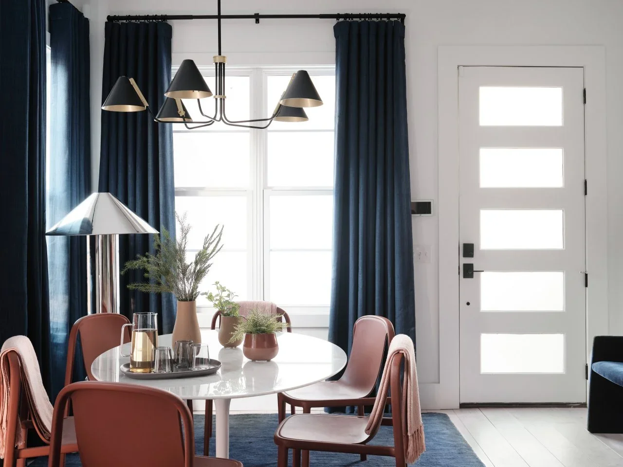 Floor-length navy curtains and a blue rug frame a white round dining table with pink chairs beside a white entrance door.