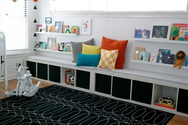 A pale nursery bench and book ledges sit above open cubbies with dark fabric bins beside a black rug with thin pale loops and colored cushions
