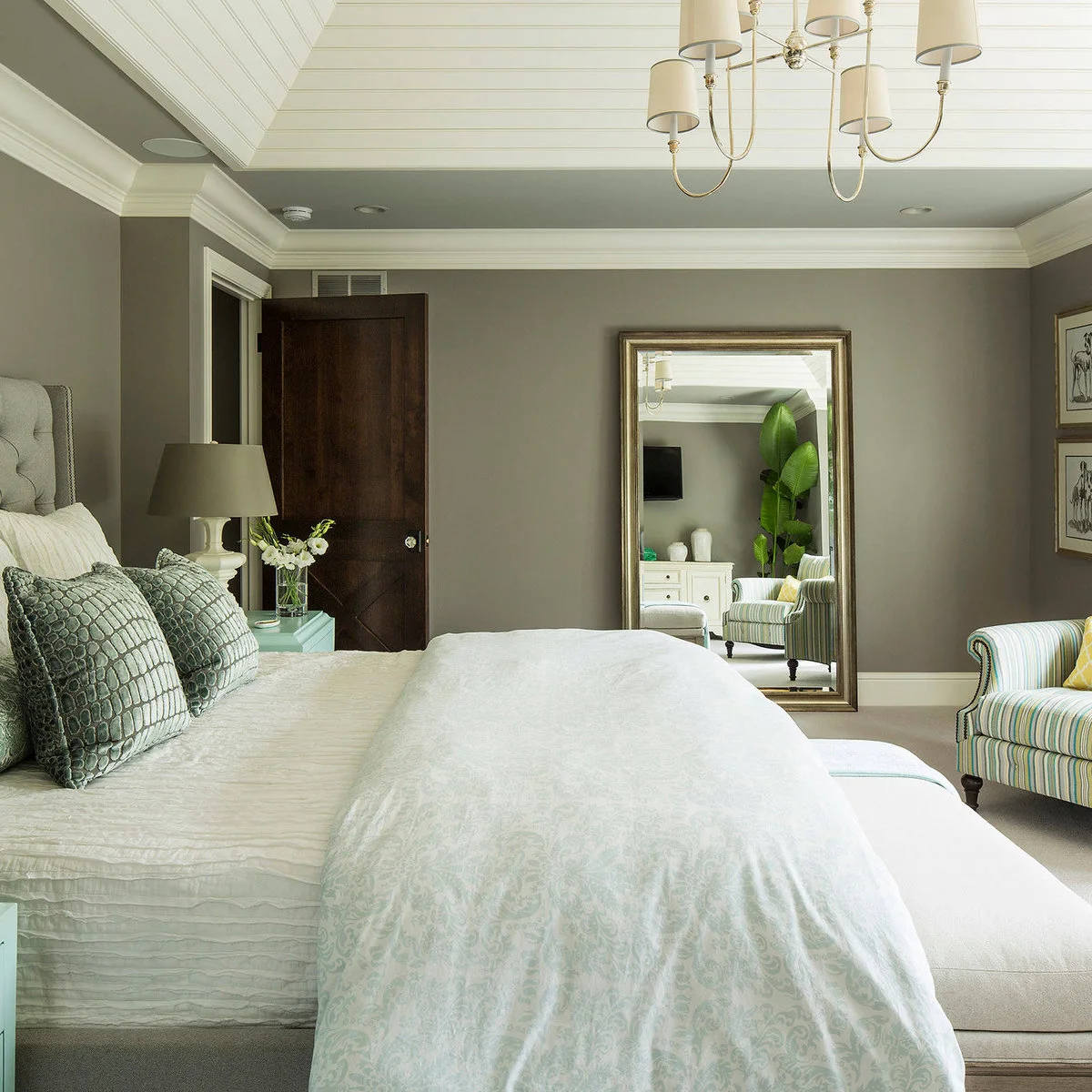 A tall framed mirror stands across from a pale bed in a gray bedroom with a striped chair and mint bedside chest.