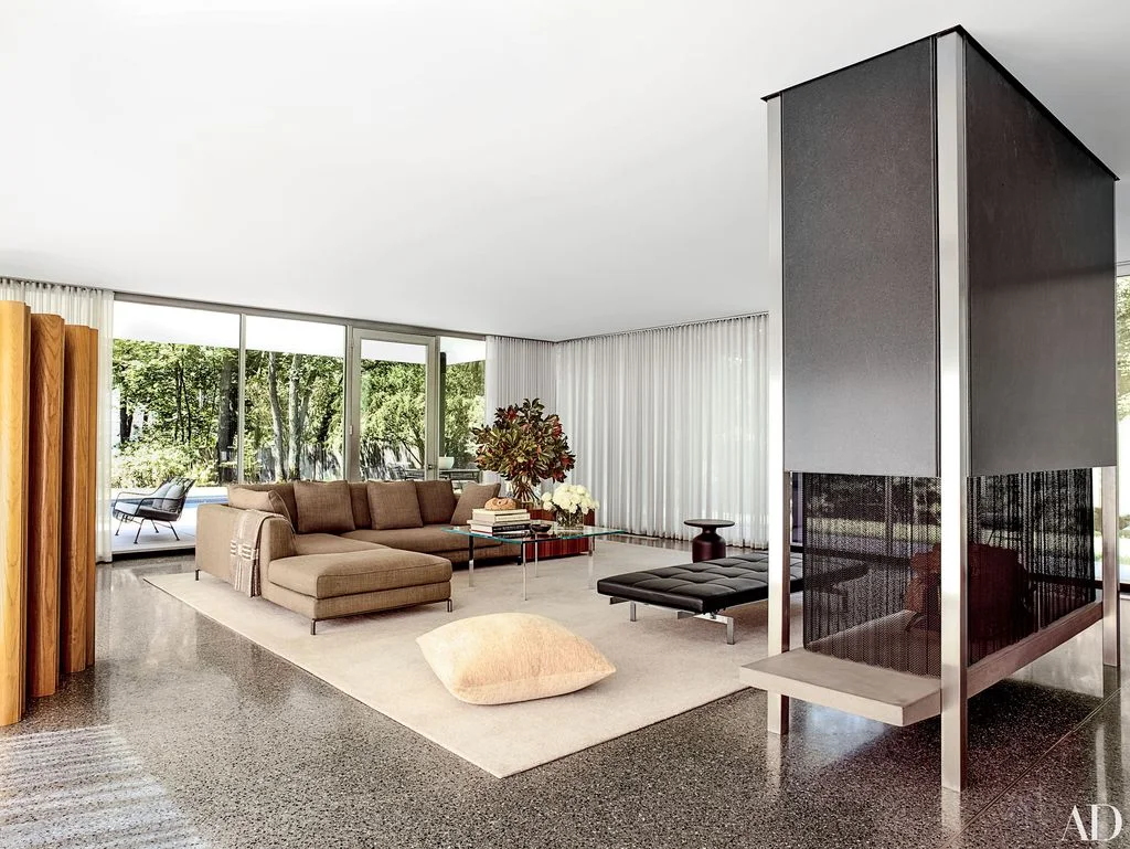 A brown sectional sofa sits on a broad light rug near large windows, pale curtains and a tall dark freestanding fireplace with screened lower sections